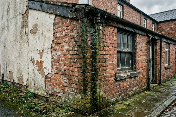 A brick wall corner with cracked render and a dark algae streak running down from a damaged, overflowing gutter